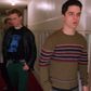 A young man in a brown knit sweater with red and navy stripes walks down a hallway, with two other men behind him wearing casual streetwear.