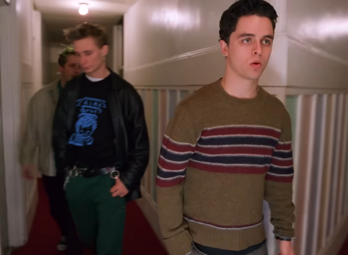 A young man in a brown knit sweater with red and navy stripes walks down a hallway, with two other men behind him wearing casual streetwear.