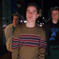 Three young men standing on a city street at night. The central figure wears a brown knit sweater with red, navy, and white horizontal stripes, while the figure on the right wears a black graphic t-shirt under a black leather jacket.