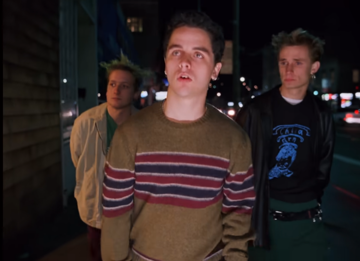Three young men standing on a city street at night. The central figure wears a brown knit sweater with red, navy, and white horizontal stripes, while the figure on the right wears a black graphic t-shirt under a black leather jacket.