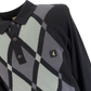 A close-up of a long-sleeve knit polo shirt featuring a diamond argyle pattern in black, gray, and pale sage green, with a small gold logo patch on the chest.