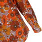 A close-up of a long-sleeve blouse featuring a bold 1970s floral pattern in orange, rust brown, mustard yellow, and dusty pink on a white background.